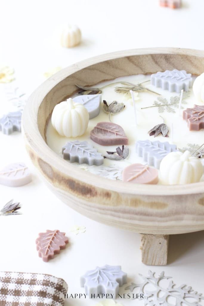 A wooden dough bowl candle filled with white wax, decorated with pastel wax leaves and pumpkins, dried flowers scattered around, and a brown gingham cloth partially visible in the corner—perfect for a cozy DIY touch.