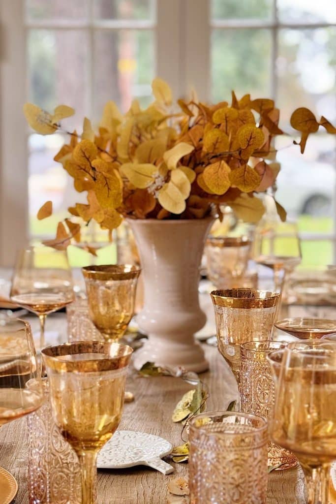 A dining table set with ornate amber glassware, gold-rimmed glasses, a white vase of autumn leaves, and a handmade candle. Natural light filters through large windows in the background.