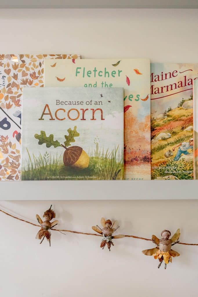 A shelf displays children's books, including "Because of an Acorn," behind a garland decorated with acorns, whimsical figures made from acorns and leaves, and a handmade Dough Bowl Candle DIY for a cozy touch.
