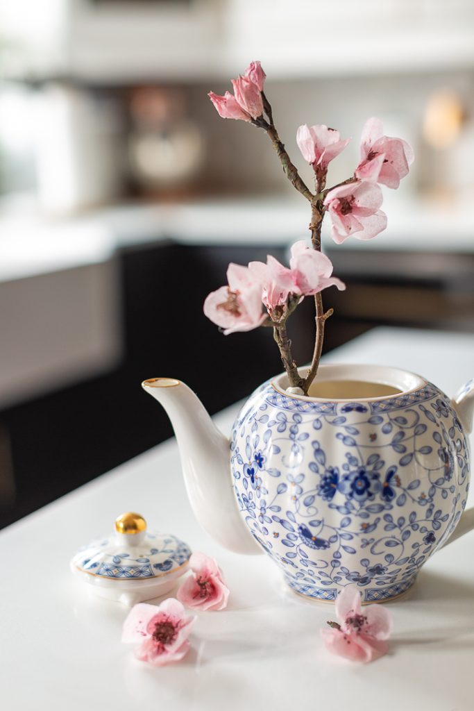 A white teapot with blue floral patterns holds a branch of pink cherry blossoms; the lid rests beside it on a white surface with scattered blossoms, offering charming inspiration for paper craft ideas.