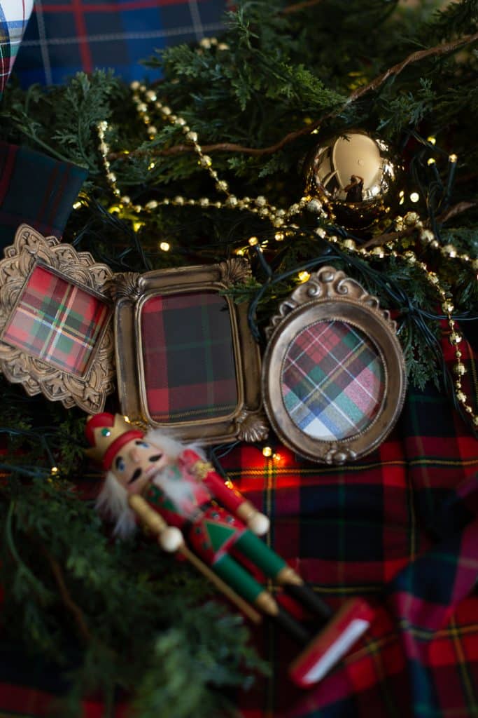 Christmas decor with tartan cloth, empty plaid-patterned picture frames, a gold ornament, string lights, gold beads, greenery, and a festive nutcracker figurine in red, green, and gold lying at the bottom alongside vintage style ornaments.